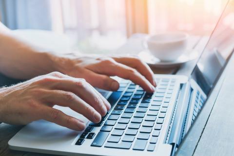 Close-up of a person's hands typing on a laptop keyboard with a white cup of coffee nearby, set on a wooden table bathed in warm sunlight filtering through a window.